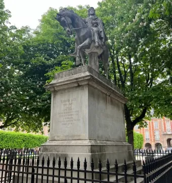 Equestrian statue of Louis XIII on a tall stone pedestal behind a black fence, with trees and a brick building in the background.