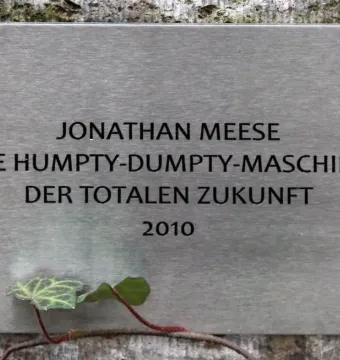 Brushed metal plaque for Jonathan Meese’s Humpty-Dumpty-Maschine mounted on a textured wall with a small ivy vine nearby.