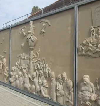 Detail of Ludwigs Erbe showing a sculptural relief with a central column and crowded figures on a beige wall.