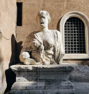 Madama Lucrezia, a weathered white stone bust of a woman on a pedestal beside a tan wall with windows.