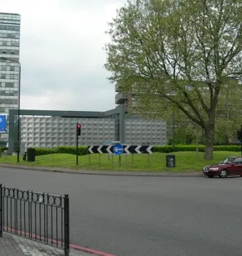Michael Faraday Memorial on a grassy roundabout island beside roads and traffic lights, with buildings and trees in the background.