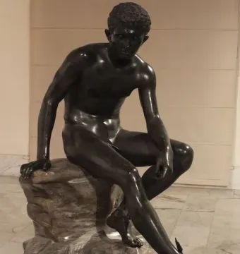 Dark bronze seated Hermes sculpture on a light-colored rock base, leaning forward with winged ankles visible.