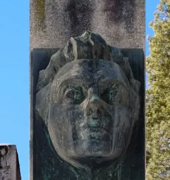 Weathered bronze bust of Julius Fučík set into a rough stone stele, with verdigris against a blue sky.