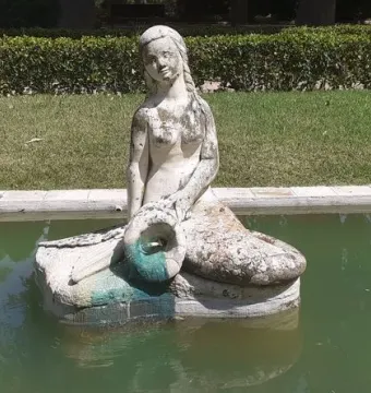Weathered stone mermaid statue in a rectangular pool with murky green water, holding a turquoise object in her lap.