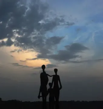 Silhouette of the Monument to the Victims of Fascism showing two adults and a child against a twilight sky, with water and trees behind it.