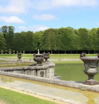 Neptune Fountain at Versailles with a large basin, decorative stone urns, and a trimmed tree hedge under a blue sky.