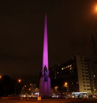 The Obelisk of Montevideo lit in purple at night, with buildings and traffic around it.