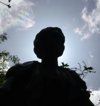 Dark silhouetted bust of the Pankhurst Memorial against a bright sky with wispy clouds and tree branches.