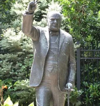 Bronze Winston Churchill statue standing outdoors, raising his right hand in a V-sign and holding a walking stick.