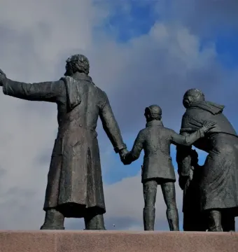 Auswandererdenkmal sculpture of three dark metallic figures standing together on a stone pedestal against a blue sky.