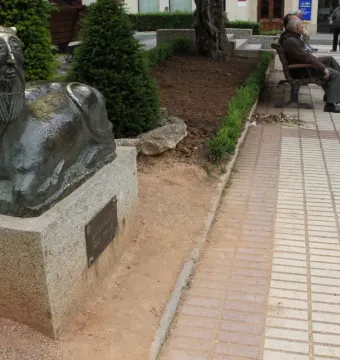 Bicha of Balazote sculpture on a stone plinth in a park, with a reclining animal body and human-like bearded head.
