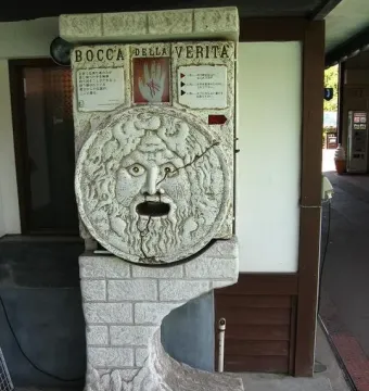 Bocca della Verità stone face replica with wide-open eyes and a gaping mouth, shown on a textured pedestal with a Japanese sign above it.