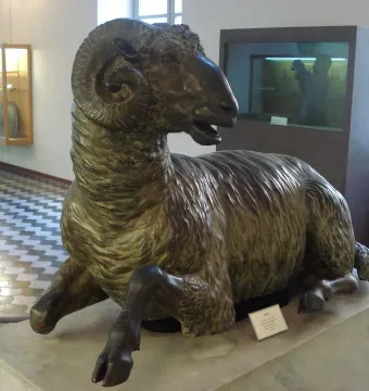 Bronze ram sculpture with curled horns resting on a stone pedestal in a museum gallery.