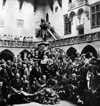 Copernicus Monument on a pedestal beside a large floral wreath, surrounded by a crowd in a grand hall.