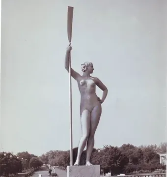 Girl with an Oar sculpture standing on a tiered pedestal in a park fountain, holding a tall oar upright.