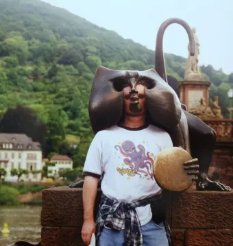 A man poses inside the face opening of the Heidelberg Bridge Monkey sculpture, with a bronze monkey holding a disc on a brick wall behind him.