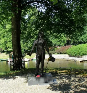 Bronze Pushkin statue on a stone plinth beside a canal, with flowers at the base and a large tree overhead.