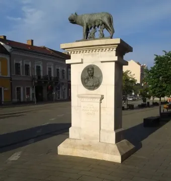 Capitoline Wolf sculpture on a stone pedestal with a bronze medallion and Latin inscription below.