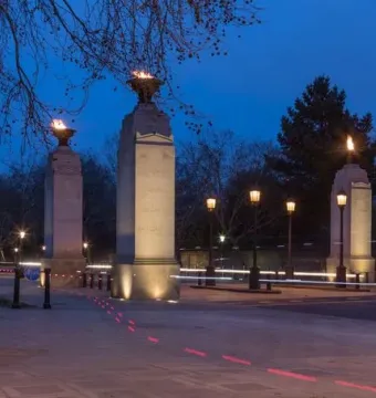 Commonwealth Memorial Gates at dusk, with a domed pavilion, four lit stone pillars topped by flames, and bare trees against a blue sky.