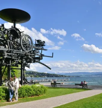 Heureka sculpture standing on a grassy lawn beside Lake Zurich, with mountains in the distance.
