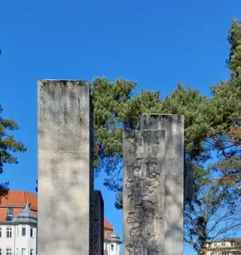 Tall stone steles of the Julius Fučík Memorial stand in a snowy park with evergreen trees and nearby buildings.