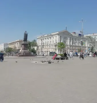 The Monument to Dimitry of Rostov stands on a tiered pedestal in a city square, with pedestrians, pigeons, and buildings in the background.