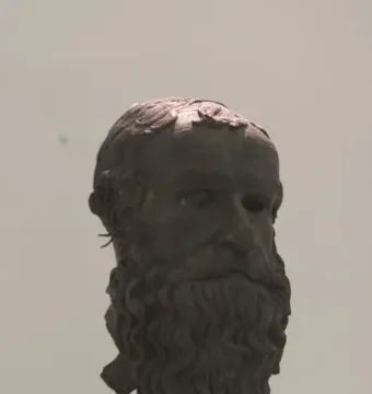 Bronze head of the Porticello Philosopher with a curly beard and worn surface on a dark stand.