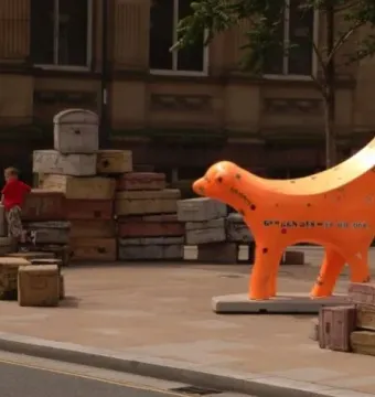 Superlambanana sculpture beside stacks of old suitcases on a paved outdoor area, with a child in the background.
