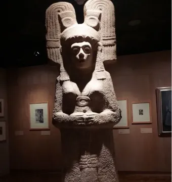 The Young Woman of Amajac sculpture displayed on a white pedestal in a dim museum hall.