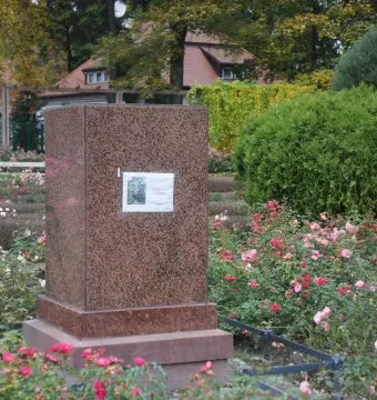 Brown granite base of The Dancing Girl monument with a small placard, surrounded by flowering bushes and trees.