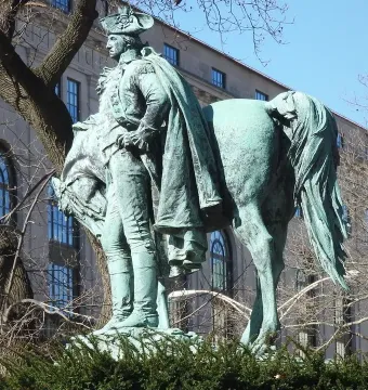 Verdigris-patinated George Washington sculpture beside a horse, seen from a low angle with bare branches and a building behind it.