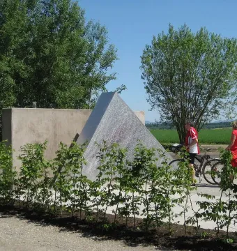 Hailfingen-Tailfingen Memorial beside a concrete wall, with two people and bicycles nearby, shrubs lining a gravel path, and fields and trees in the background.