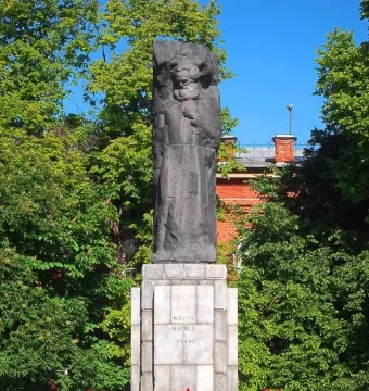 Karl Marx monument with a dark stone figure on a tiered base, framed by green trees and a brick building.