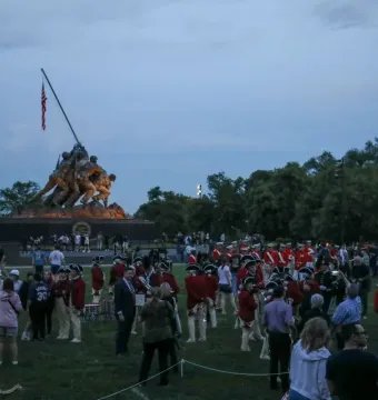 The Marine Corps War Memorial stands illuminated at dusk with bronze figures raising a flag, while a crowd gathers on the lawn below.