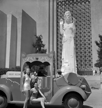 Pacifica statue of a woman with raised hands and an elaborate headdress stands before a star-patterned wall, with two women posing beside a sightseeing vehicle.
