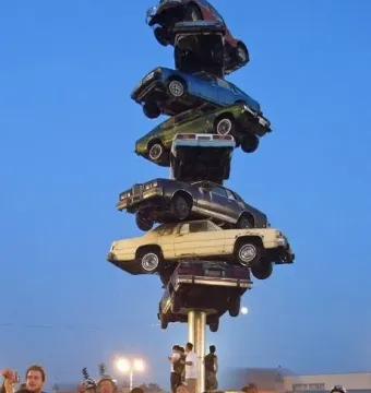 The Spindle sculpture made of vertically stacked cars with people and bicycles gathered around it at dusk.