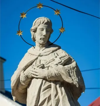 Sandstone statue of St. John of Nepomuk with a halo of stars against a blue sky.