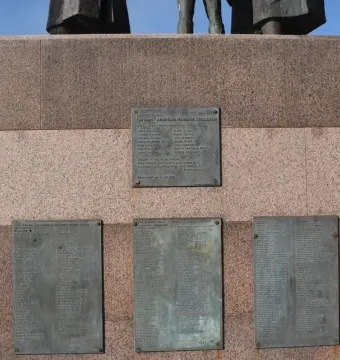 Bronze name plaques on the granite base of the Auswandererdenkmal, with the lower legs of the figures visible above.