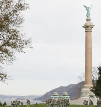 Battle Monument at West Point with soldiers marching past it beside the Hudson River.
