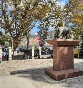 Capitoline Wolf sculpture with Romulus and Remus on a tall red-stone pedestal in an urban square.