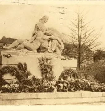 Stone war memorial with two sculpted figures embracing on a pedestal, surrounded by wreaths and flowers.
