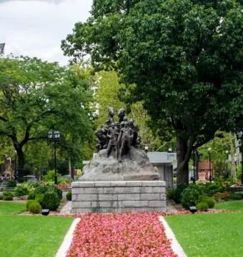 Wars of America bronze sculpture on a stone base in a green park with trees and a flower-lined path.