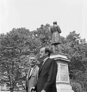 Two men in suits walk in front of the Johan Ludvig Runeberg statue in a park.