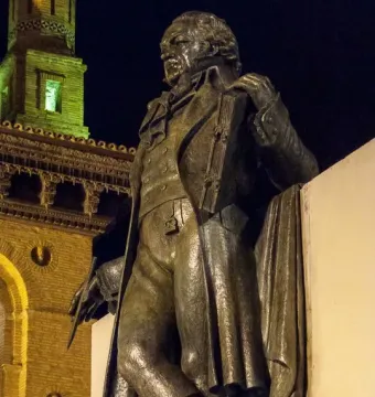Goya Memorial bronze sculpture of a man in a period coat holding a paintbrush, with a lit stone building behind it at night.