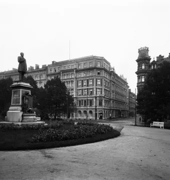 Johan Ludvig Runeberg statue on a large stone pedestal along a park pathway, with historic buildings and trees behind it.