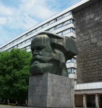 Karl Marx Monument, a large dark bust on a tall granite pedestal, with a modern building behind it.
