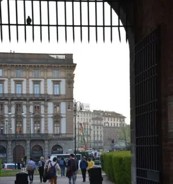 Needle, Thread and Knot sculpture seen through a dark metal gateway, with pedestrians walking along a paved path in Milan.