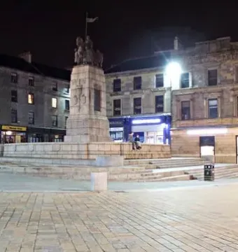 Paisley War Memorial on a stone pedestal in a lit town square at night, with surrounding buildings and streetlights.
