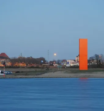 Rhine Orange sculpture standing on the far bank of a wide river with a distant townscape behind it.