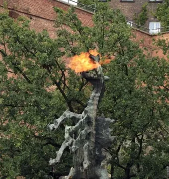 Wawel Dragon sculpture spewing a bright flame from its open mouth beside trees and a brick building